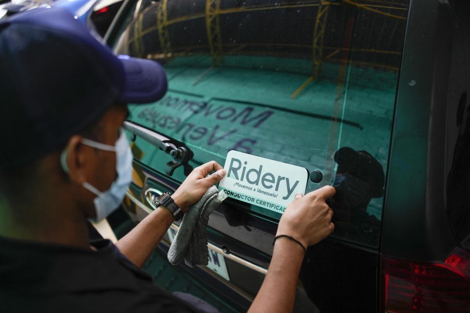 Un empleado coloca una calcomanía promocional en la ventana trasera de un vehículo en la sede de la firma Ridery en Caracas, el miércoles 4 de mayo de 2022. Ridery es una de al menos tres firmas venezolanas de taxis por app que se lanzaron durante la pandemia, y que han aprovechado un cambio de facto de monedas, del bolívar venezolano al dólar estadounidense. (AP Foto/Matias Delacroix)