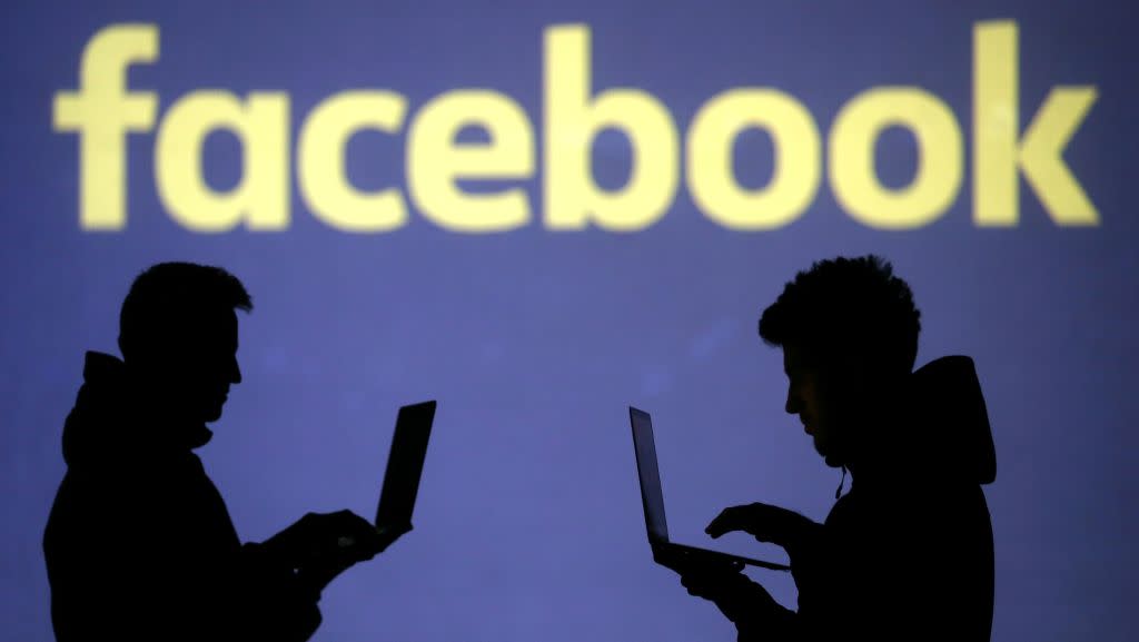 Silhouettes of laptop users are seen next to a screen projection of Facebook logo.