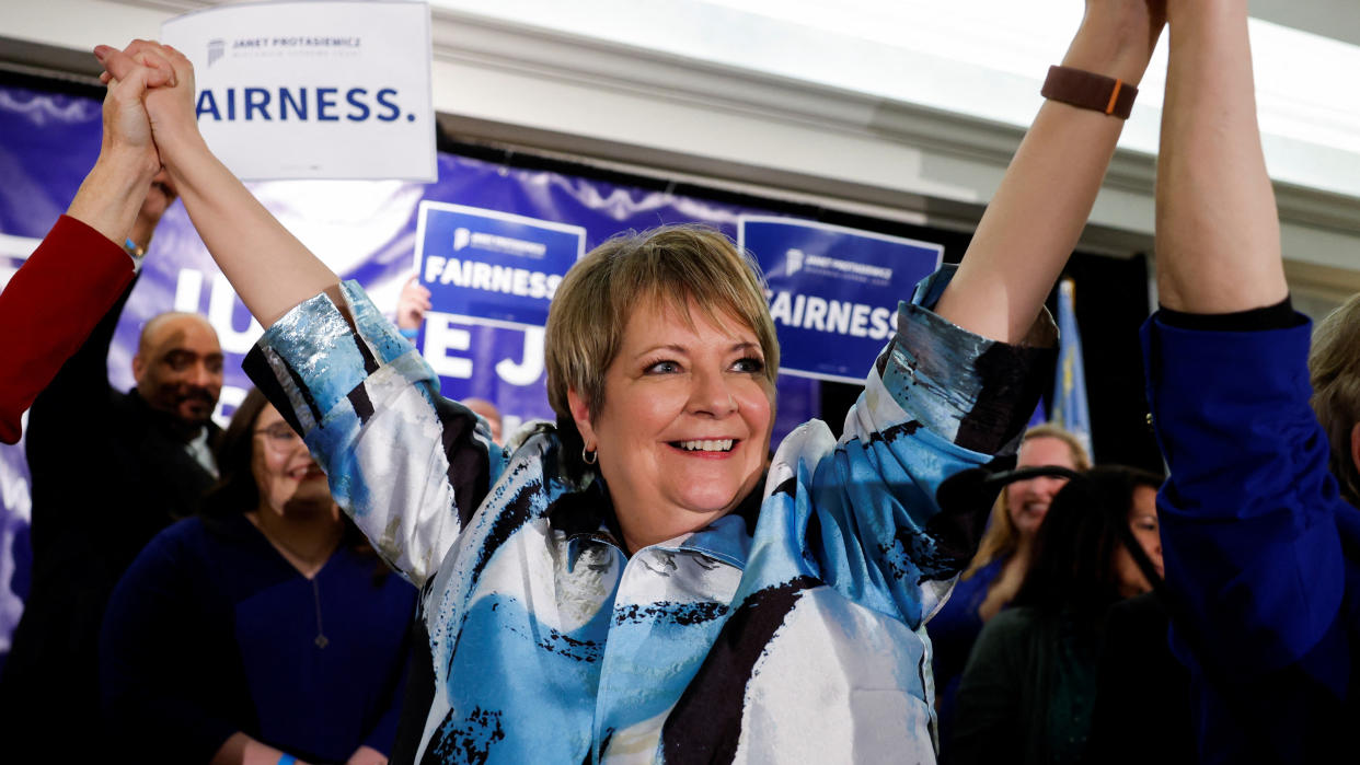 Janet Protasiewicz raises both arms in the air, and links hands with supporters, against a backdrop of posters saying: Fairness.