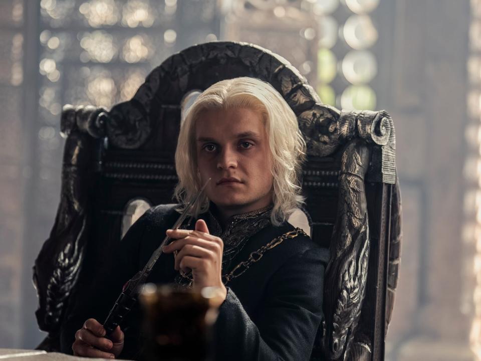 tom glynn-carney as aegon targaryen in house of the dragon. he's sitting in a high backed chair, playing with a steel dagger, and looking across a table