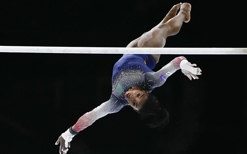 Gymnast Simone Biles is in midair during competition. A bar stretches horizontally across the image, and Biles is behind it, with her feet near the top of the image and her head toward the bottom. Her arms are out and she is intensely focused on the bar.