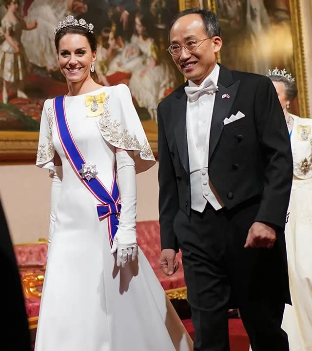 Princess Catherine walks in a long white dress with a blue sash and yellow brooch next to a man in black suit