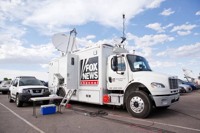 A Fox News Channel satellite truck is parked in a lot with equipment outside next to an SUV under a partly cloudy sky