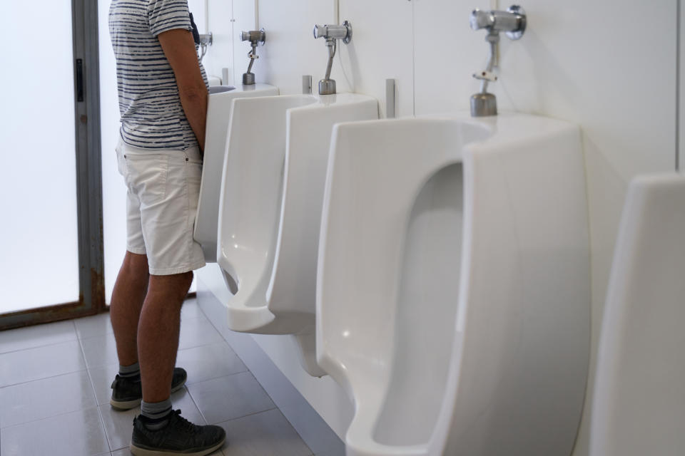 A person in a striped shirt and shorts is standing in front of a row of urinals in a bathroom