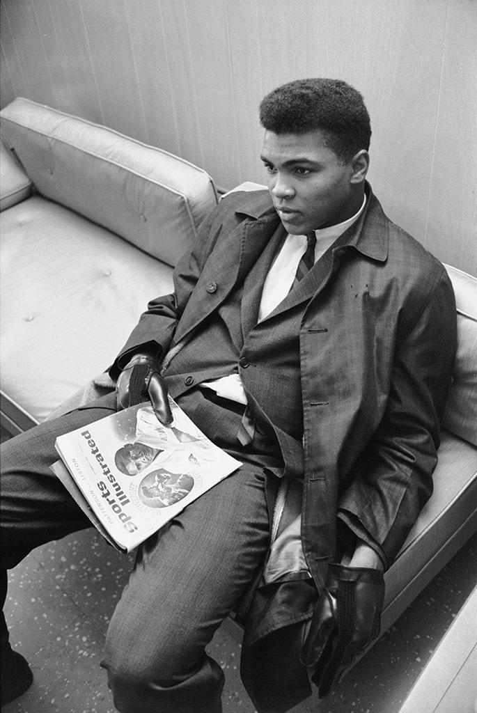 Ali (then known as Cassius Clay) sits on a couch, with a copy of Sports Illustrated on his lap.