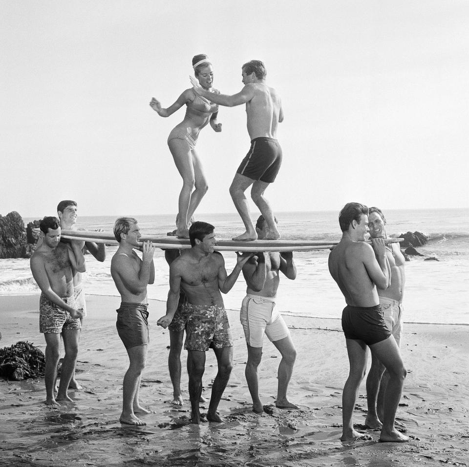 FILE - This 1965 file photo of youthful actors in a Hollywood movie amuse themselves between shooting scenes at California's Malibu Beach by staging an airborne twist exhibition on top of a surf board. For some Native Hawaiians, surfing's Olympic debut is both a celebration of a cultural touchstone invented by their ancestors, and an extension of the racial indignities seared into the history of the game and their homeland. The Summer Games in Tokyo, which kick off this month, serve as a proxy for that unresolved tension and resentment, according to the Native Hawaiians who lament that surfing and their identity have been culturally appropriated by white outsiders who now stand to benefit the most from the $10 billion industry. (AP Photo, File)