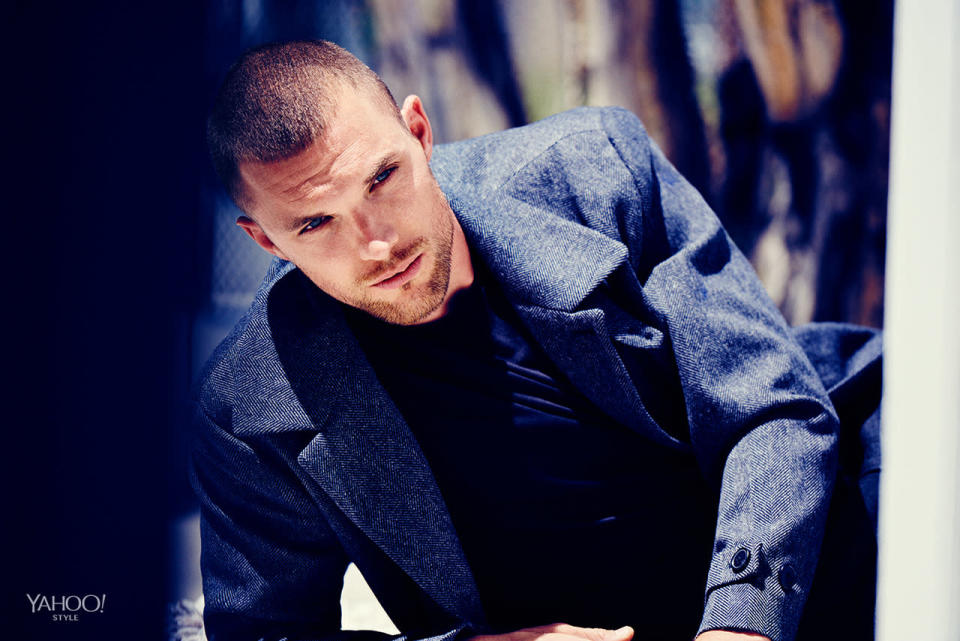 Ed Skrein kneels down in a Theory gray wool coat.