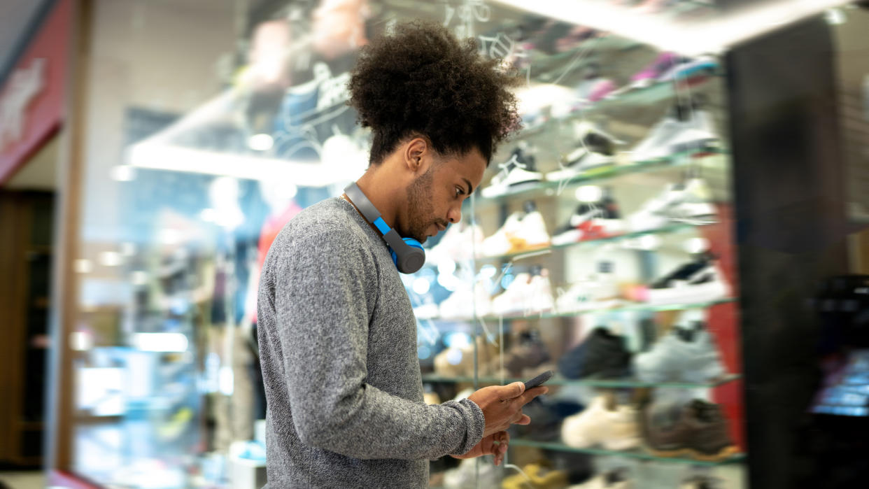  Man looks at phone while standing in front of a store window display of sports shoes. 