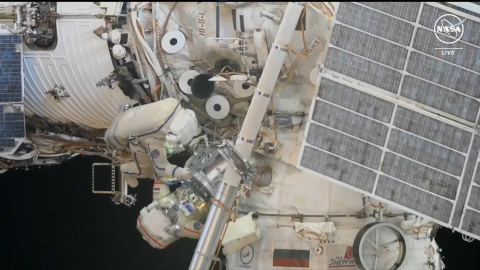 two astronauts in white spacesuits climb on white cylinders floating in space