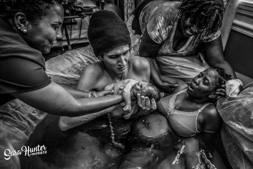 An exhausted mother leans back after delivering in a water birth while the dad holds the new baby