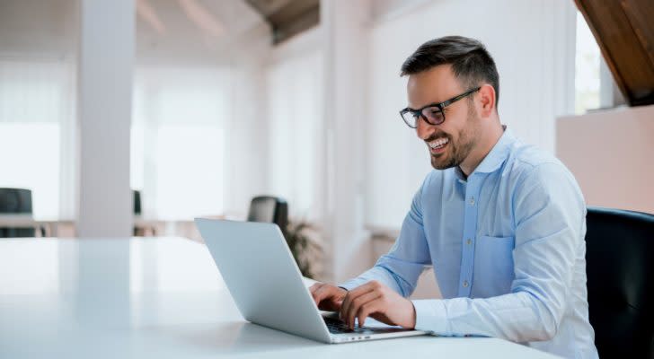 a man in a collared shirt smiles while typing on his laptop