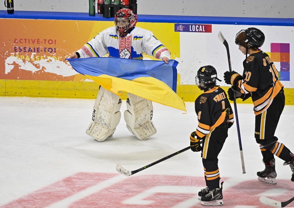 CORRECTS UKRAINE GOALIE NAME TO DMYTRO KORZH, NOT MATVII KULISH - Ukraine peewee team goalie Dmytro Korzh, left, celebrates his team's victory by holding the Ukraine flag as Boston Junior Bruins Patrick Fennell and Brendan O'Toole skate by at the end of a hockey game, Saturday, Feb, 11, 2023, in Quebec City. (Jacques Boissinot/The Canadian Press via AP)