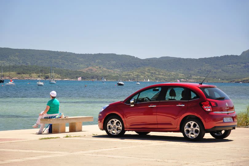 A person sitting on a bench beside a beach with a red car behind them