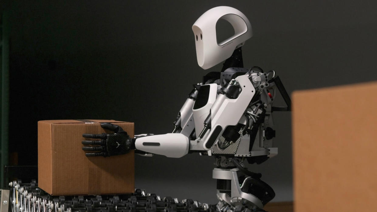  A white and black humanoid robot lifts a small cardboard box. 