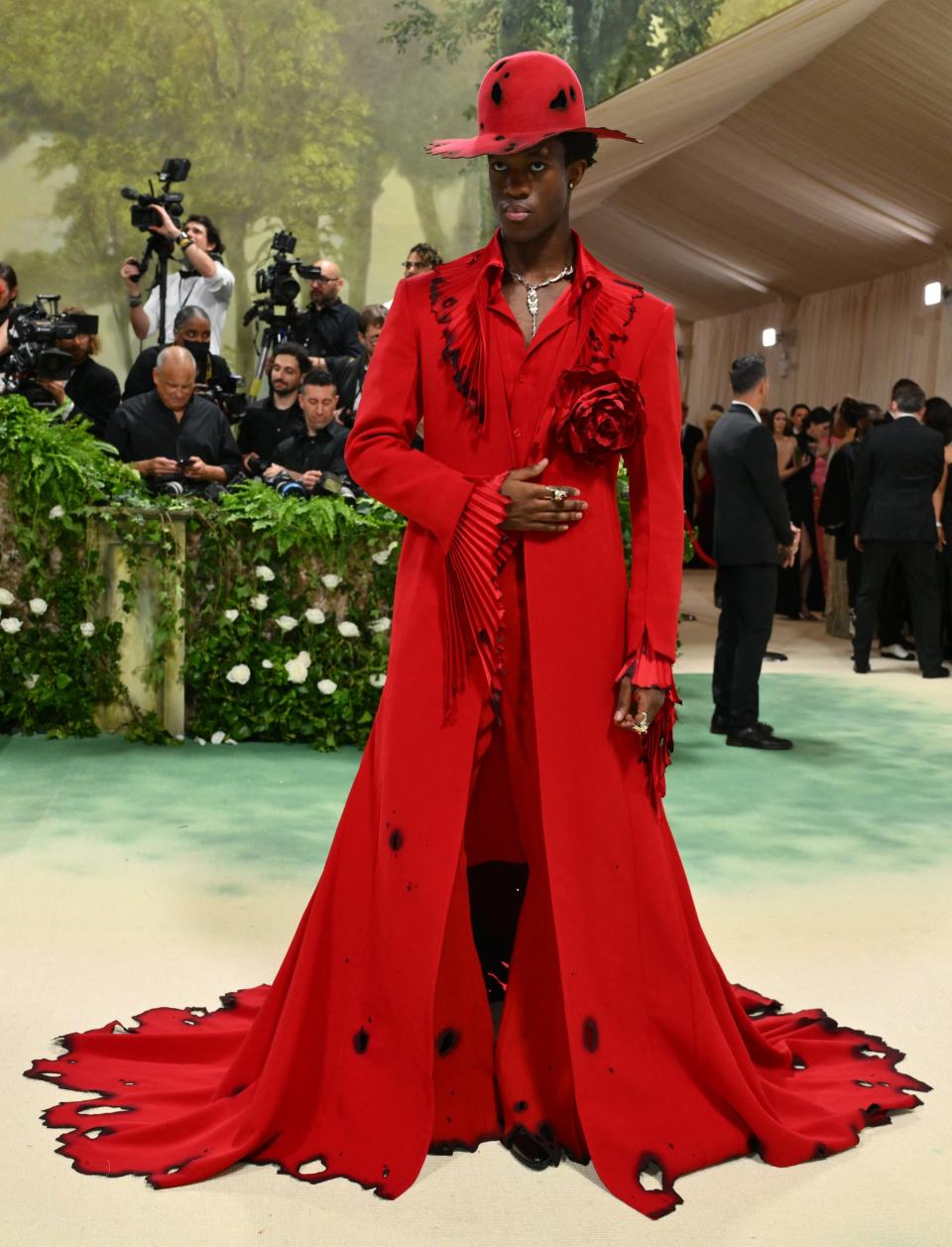Person in a dramatic red outfit with hat and train, posing with a rose at an event. Photographers in background