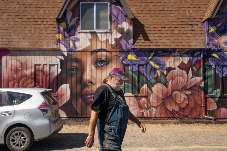 A man wearing overalls walks by a building with a face painted on it.