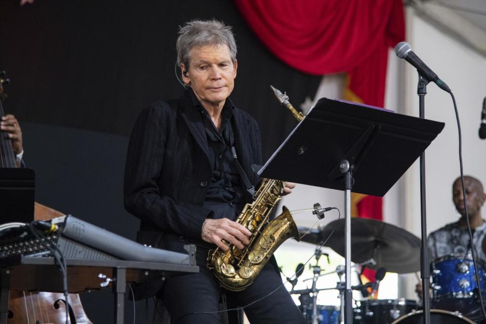 A man with gray hair in a dark outfit standing on a stage holding a saxophone behind a music stand