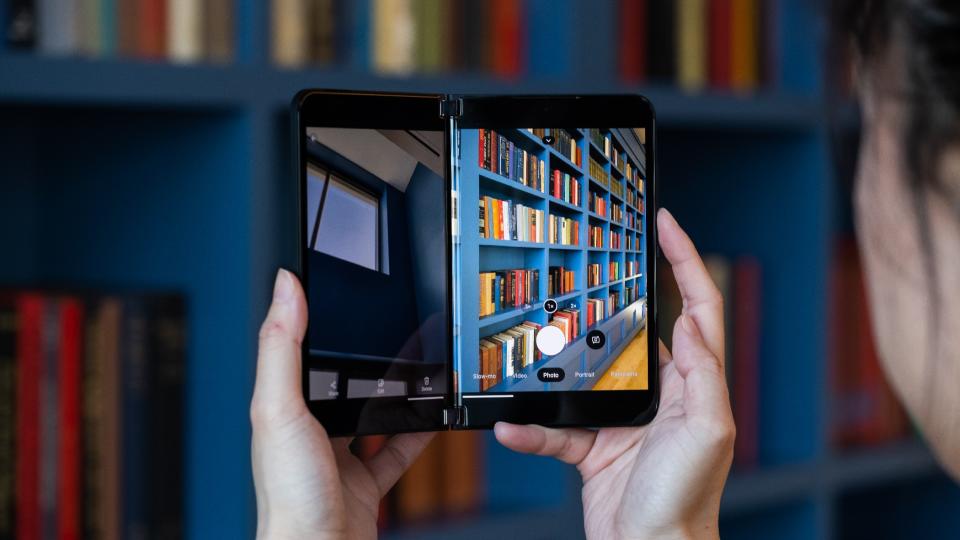 A person holding up the Microsoft Surface Duo 2 to take a photo of a bookshelf. The screen on the right shows the viewfinder while the screen on the left displays a picture taken earlier. 