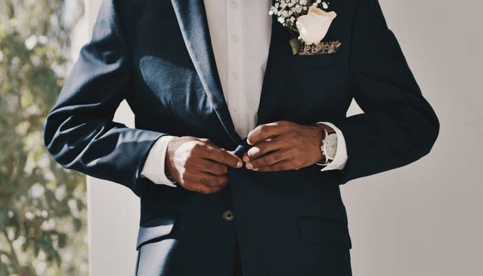 A man buttoning his suit jacket, wearing a boutonniere with a white flower and baby's breath. His white dress shirt is visible under the jacket