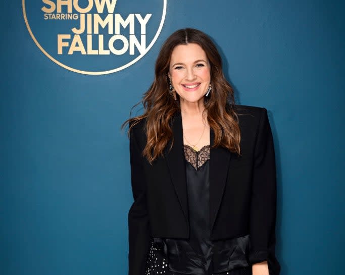 Drew Barrymore smiles in an elegant blazer outfit with a sheer dotted skirt, standing in front of a "The Tonight Show Starring Jimmy Fallon" sign