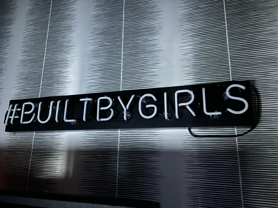 <p>A photo from the iPhone 14 showing an unlit neon sign making out the words "Built By Girl" on a striped wallpaper.</p>
