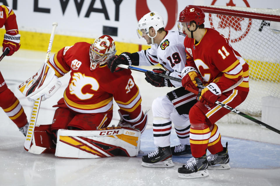 Chicago Blackhawks' Jonathan Toews, center, is checked into Calgary Flames goalie David Rittich, left, by Mikael Backlund during the third period of an NHL hockey game Tuesday, Dec. 31, 2019, in Calgary, Alberta. (Jeff McIntosh/The Canadian Press via AP)