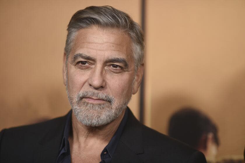 George Clooney wears a black shirt and blazer and stands in front of a golden backdrop