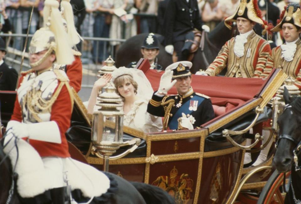 princess diana and prince charles sit in an open carriage and wave and smile at crowds, she wears a veil and wedding dress, he wears a military uniform including a hat and white gloves