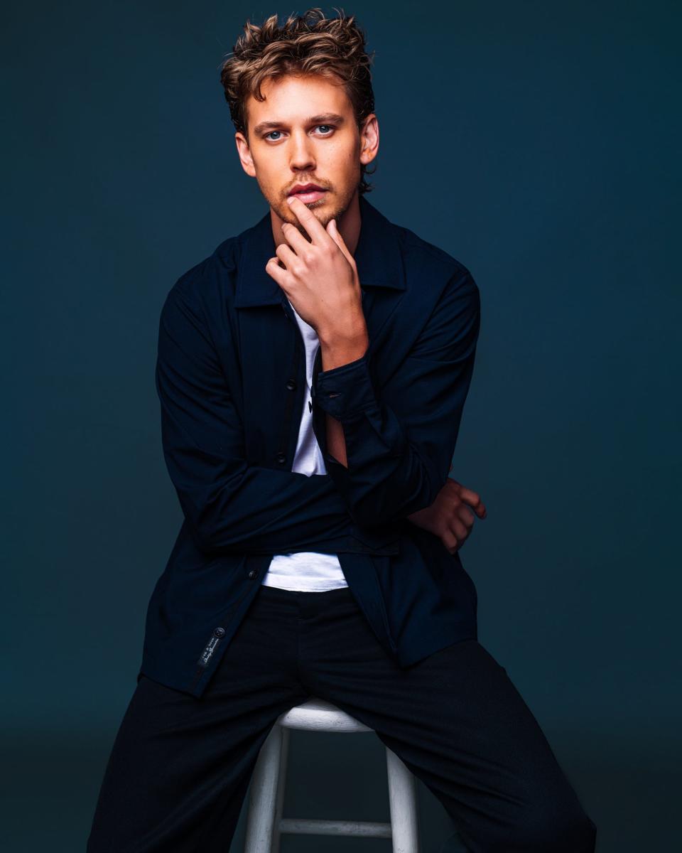 Austin Butler poses in a casual outfit, wearing a white t-shirt and dark jacket. He is sitting on a stool, resting his chin on his hand while looking at the camera