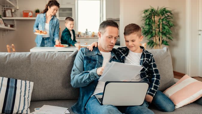 Father Works From Home While His Younger Son Makes Him Company While Mum With Other Son Sets Lunch in the Background.