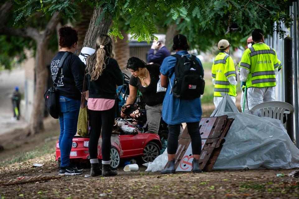 Several people stand near a red toy car and a white tarp covering under the shade of trees