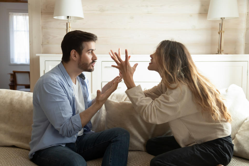 A man and a woman are sitting on a couch, engaged in an intense argument, gesturing with their hands. Two lamps and a window are in the background