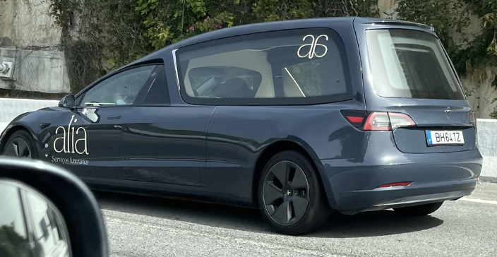 Blue hearse with "alia" business logos on the side and rear window, seen driving down a road