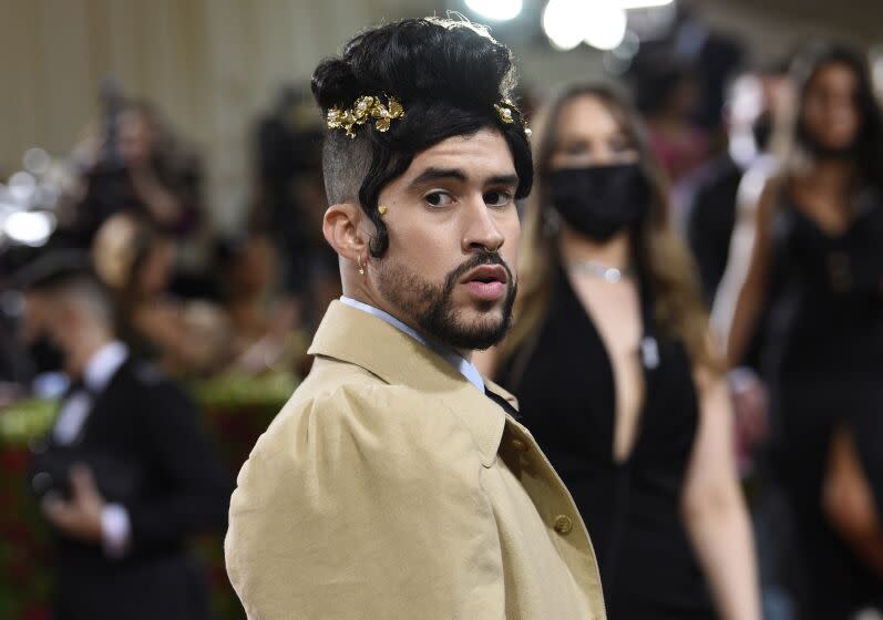 A man wearing his black hair in a bun atop his head and posing in a beige coat