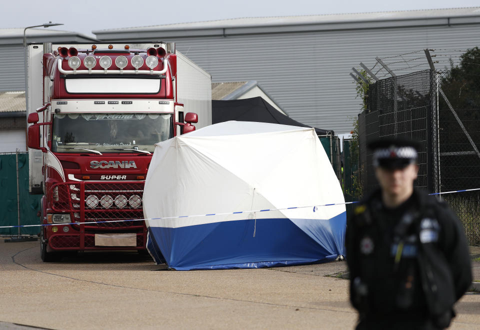 A police officer stands guard in front of a truck, in rear, that was found to contain a large number of dead bodies, in Thurrock, South England, early Wednesday Oct. 23, 2019. Police in southeastern England said that 39 people were found dead Wednesday inside a truck container believed to have come from Bulgaria. (AP Photo/Alastair Grant)