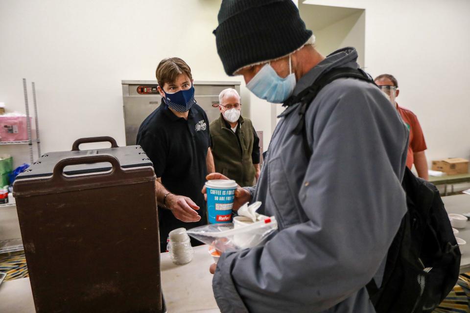 Ford Motor Co. CEO Jim Farley left, hands a cup of coffee to Shane Baertsch, 47, who is staying at the St. John's Community Center, as he grabs his breakfast at the Pope Francis Center at TCF Center in downtown Detroit on March 6, 2021. Fr. Tim McCabe, S.J., looks on in the background.