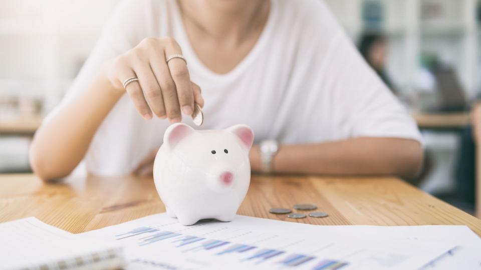 Woman sits at table with investment charts on pieces of paper, she's putting coins into a piggy bank. 