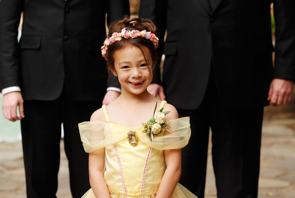 Lily in a floral headband and princess dress smiles, standing in front of two adults in black suits.