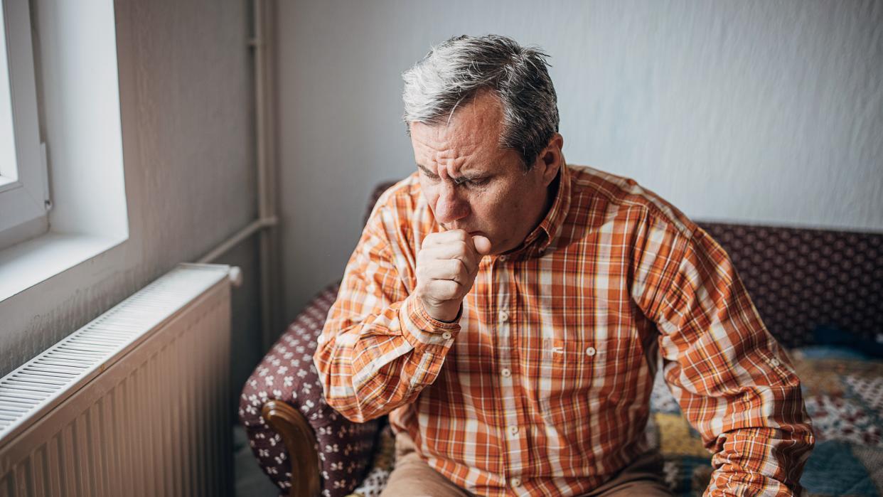  Photo of an older man sitting on a couch indoors and coughing into a closed fist. 