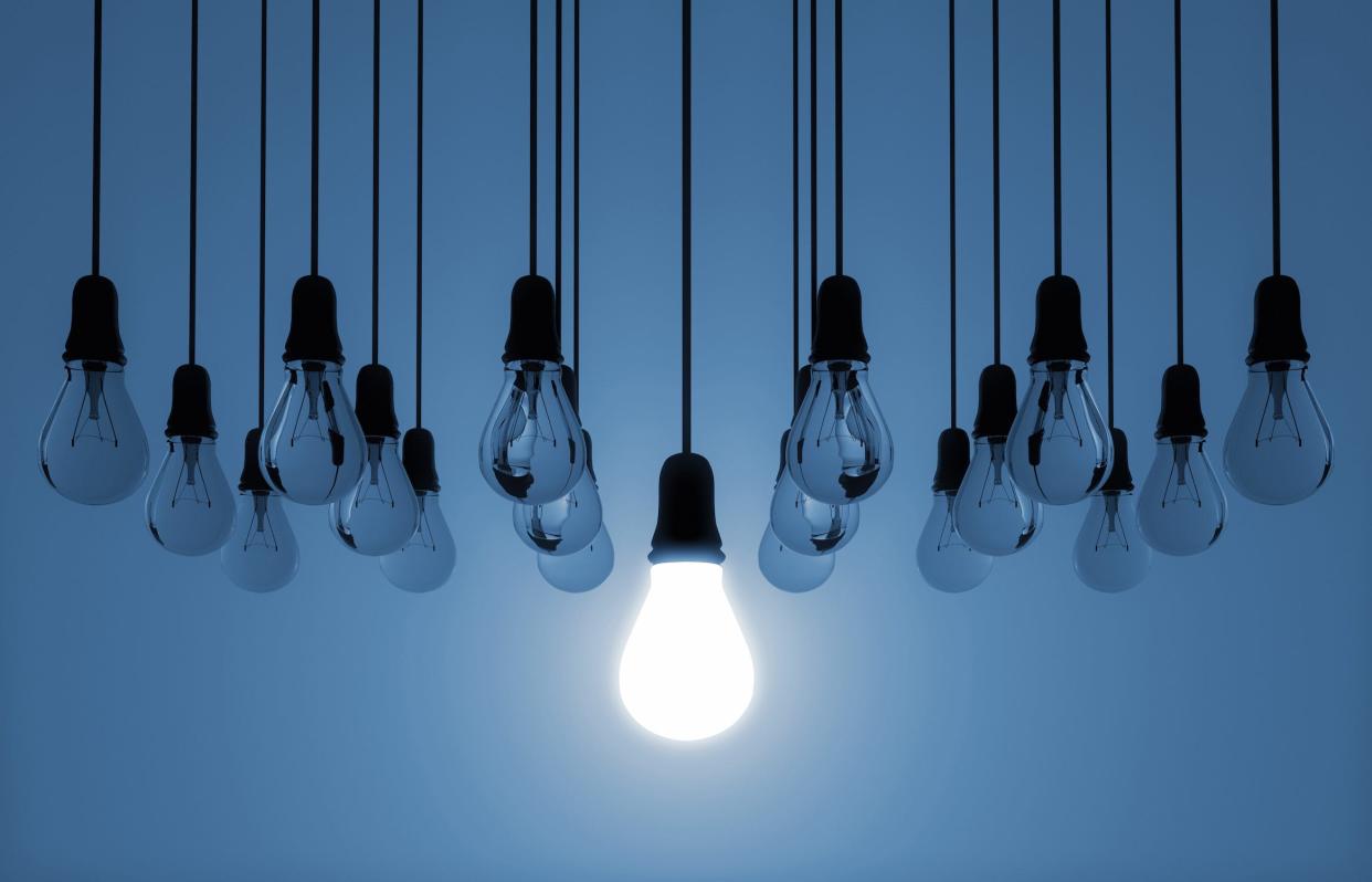 array of lightbulbs hanging dark unlit against a blue background with one lit bulb hanging lower in the center