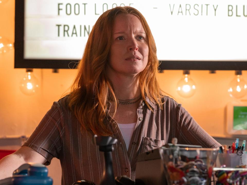 Adult Van, in a brown shirt and silver jewelry, stands behind the counter of her movie rental shop in this still from "Yellowjackets" season two.