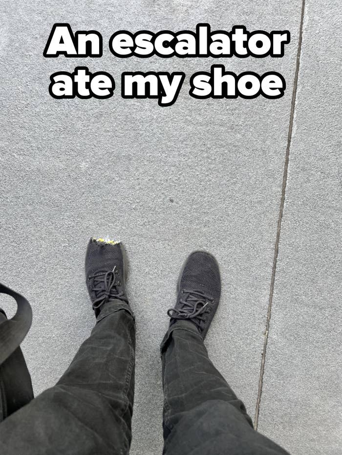 Person wearing black sneakers with one sneaker's toe area visibly worn out, standing on a concrete surface. Only their legs and feet are visible