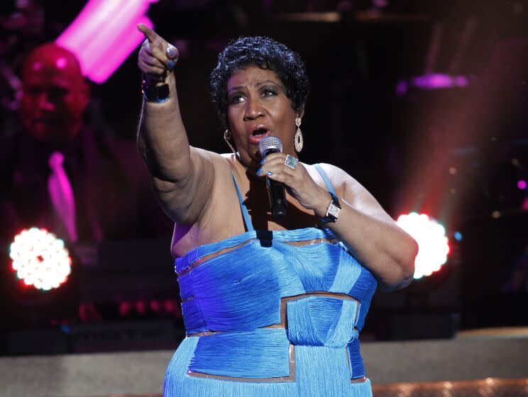 Aretha Franklin is pointing while holding a microphone to her mouth as she sings on stage, wearing a blue dress