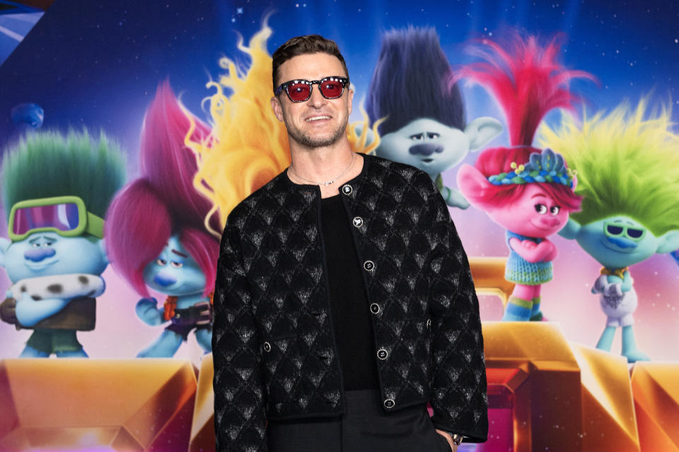 Justin Timberlake on the red carpet in front of a "Trolls World Tour" backdrop featuring animated characters. He wears a patterned jacket and sunglasses