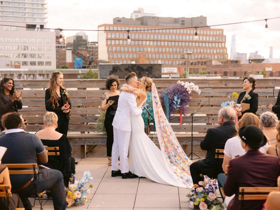 A bride in a white suit and a bride in a white dress and rainbow veil kiss at their wedding altar.