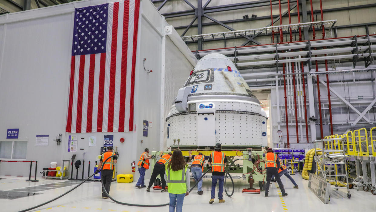  Technicians surround a white space capsule in a large room, with an american flag on one wall. 