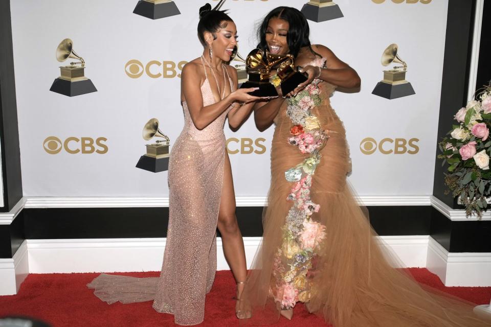 doja cat and sza each hold a grammy trophy and smile while standing together, both women wear dresses