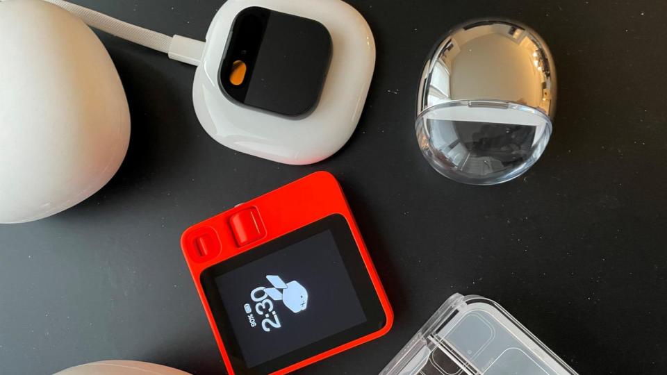 PHOTO: A Rabbit R1 and a Humane AI Pin stand on a table next to their charging and carrying cases, April 29, 2024. (Courtesy of Jonah Cohn)