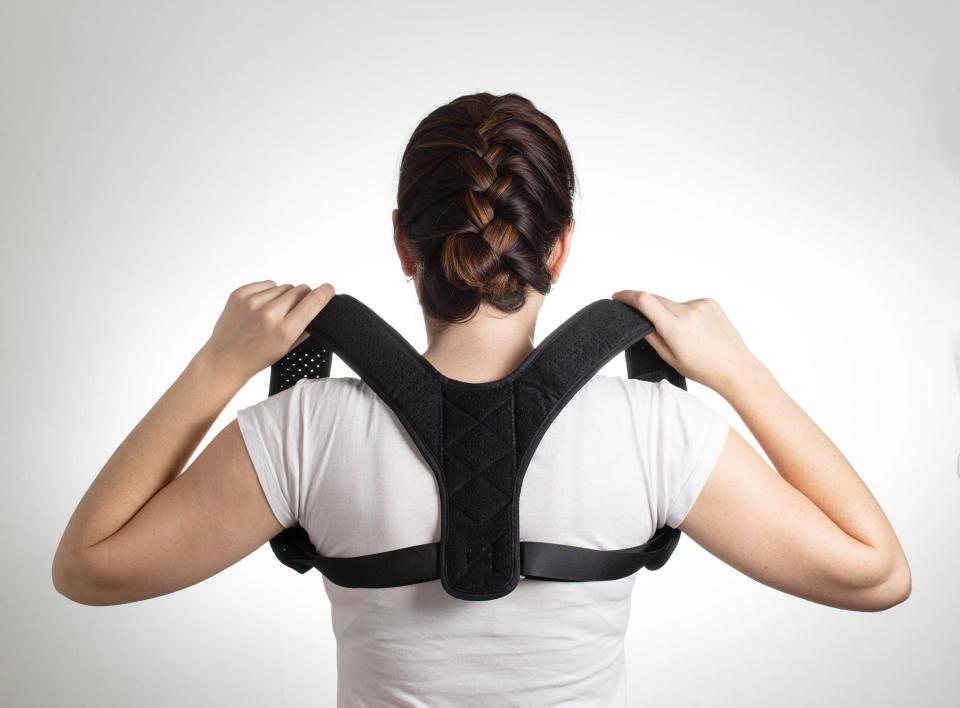 A woman in a white t-shirt and a posture-correcting harness stands with her back to the viewer