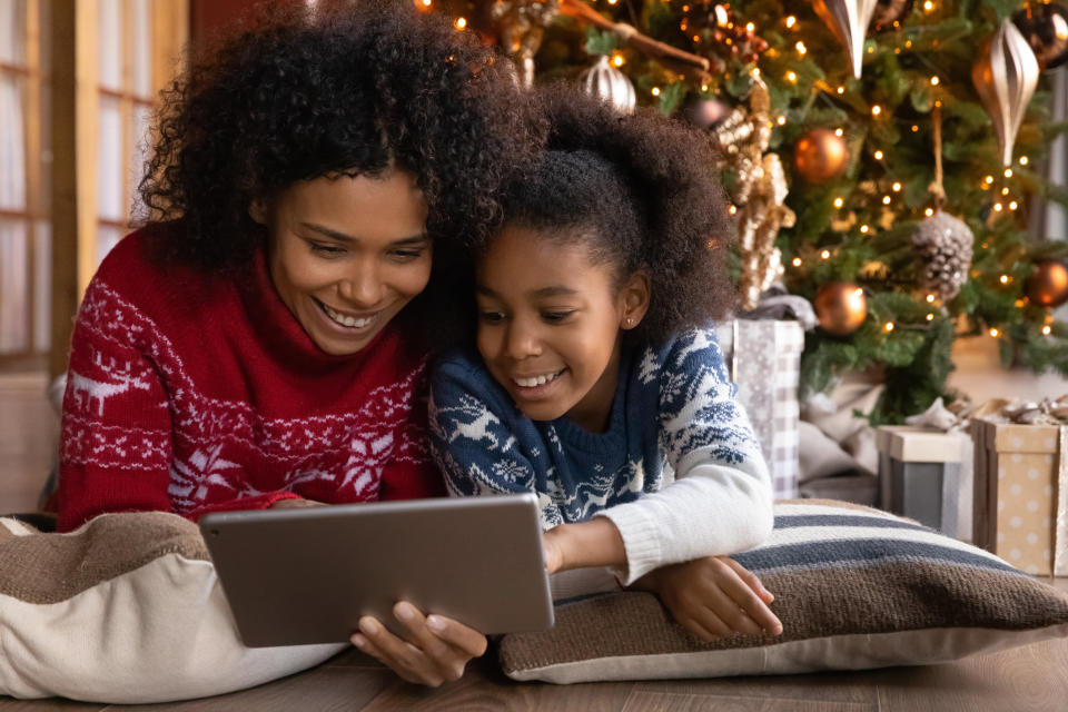 Close up happy African American woman with daughter using tablet together, lying on soft pillows near Christmas tree at home, happy family shopping online, choosing gifts, enjoying winter holiday